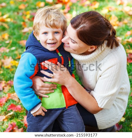 Young mum and her little kid son hugging together in autumn park on sunny day in colorful clothes.