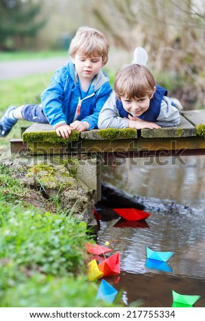 Two little brothers playing together with colorful paper boats by a river on spring or autumn day. Creative leisure with kids.