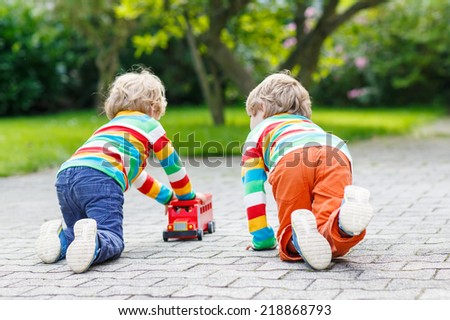 Two little friends boys in colorful clothing with stripes playing with red school bus toy in summer garden on warm sunny day. Learning to play and communicate together.