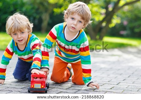 Two little siblings, kid boys in colorful clothing with stripes playing with red school bus toy in summer garden on warm sunny day. Learning to play and communicate together.