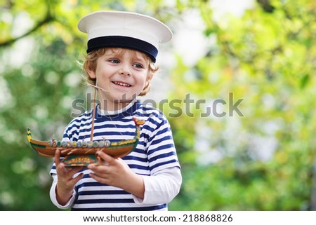 Happy little child in skipper uniform playing with toy ship against green tree summer background