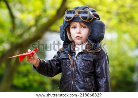 Happy kid boy in pilot helmet playing with toy airplane against green tree summer background