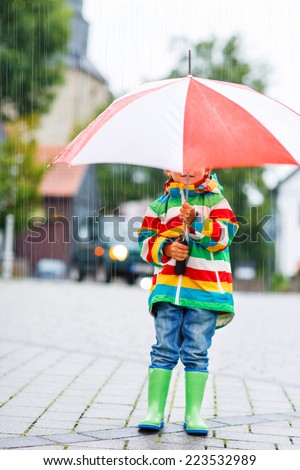 Cute little boy walking in city and hiding under red umbrella, wearing colorful rain coat and green boots outdoors at rainy day
