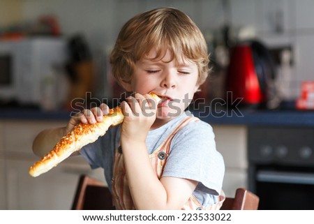 Little boy eating long loaf bread or baguette in kitchen