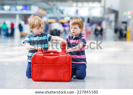 Two little sibling twins having fun and going on vacations trip with suitcase at airport, indoors. Kids packing red suitcase.