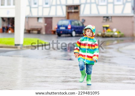 Happy smiling little boy walking in city through rain, wearing colorful rain coat and green boots outdoors at rainy day. Having fun.