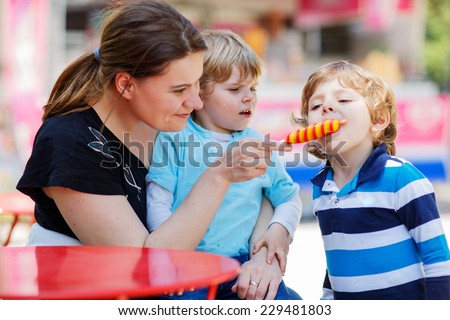 Mother feeding her little kid boys with ice cream, on warm sunny day. Happy family: mom and two sibling brothers.