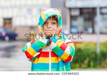 Funny smiling little boy walking in city through rain, wearing colorful rain coat and green boots outdoors at rainy day. Having fun.