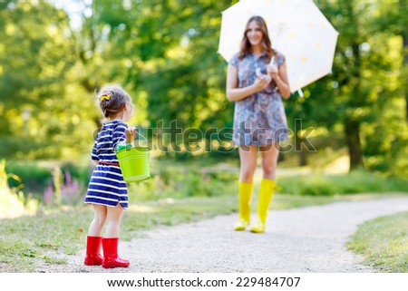 Mum and her little adorable kid daughter in rain boots, playing together in summer sunny park, on warm day.