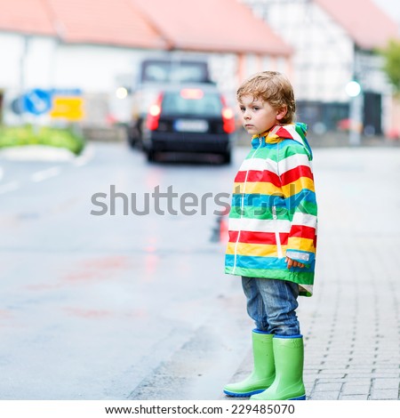 Funny cute kid boy walking in city through rain, wearing colorful rain coat and green boots outdoors at rainy day. Having fun. Square format.