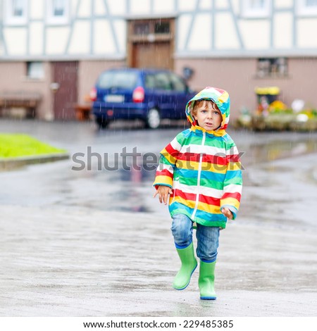 Happy smiling little boy walking in city through rain, wearing colorful rain coat and green boots outdoors at rainy day. Having fun. Square format.