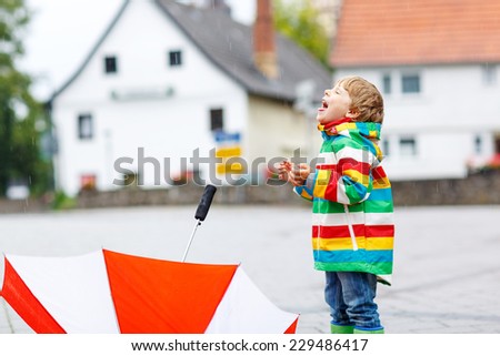 Happy smiling little boy walking in city and playing with red umbrella, wearing colorful rain coat and green boots outdoors at rainy day. Catching raindrops with mouth.