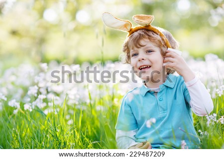 Cute happy little child celebrating Easter holiday and having fun with wearing Easter bunny ears at spring green grass and blooming apple garden, outdoors.