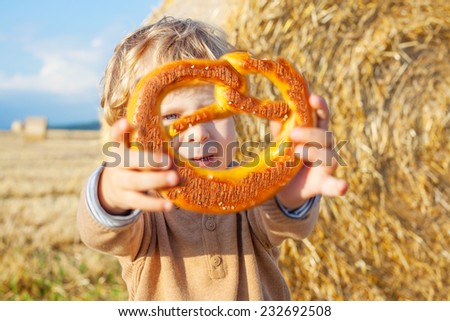 Funny cute little kid boy eating big traditional German pretzel on late summer day on wheat field with haybales.