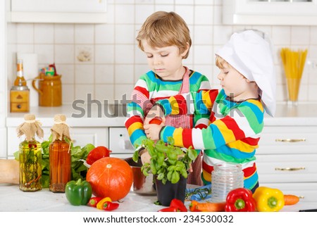 Two funny little boys cooking italian soup and meal with fresh vegetables in home\'s white kitchen. Sibling children in colorful shirts.
