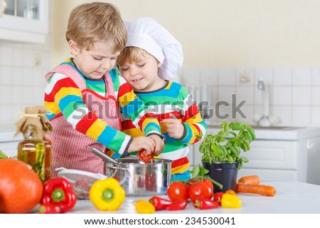 Two cute little kid boys cooking italian soup and meal with fresh vegetables in home\'s white kitchen. Sibling children in colorful shirts.