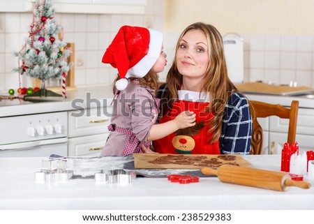 Happy mother, young woman in red santa hat baking christmas cookies together with her little daughter, cute blonde kid girl, sitting at the table in domesti? kitchen. Kid giving mum a kiss.
