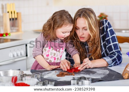 Happy mother, young woman in red santa hat baking christmas cookies together with her little daughter, cute blonde kid girl, sitting at the table in domesti? kitchen