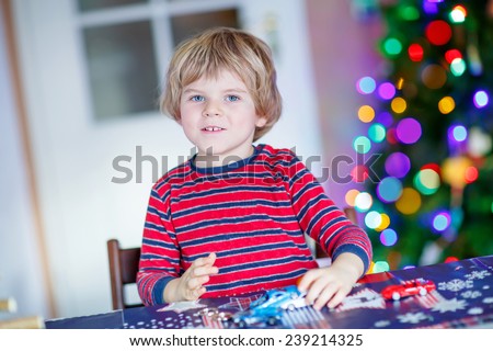 Little blond child playing with cars and toys at home, indoor. Cute happy funny boy having fun with gifts. Colorful lights on background