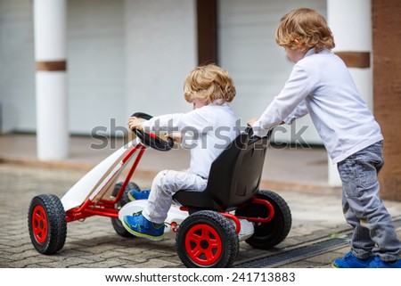 Two happy twins boys playing with toy car in summer garden, outdoors. Children having fun in domestic garden or nursery on sunny warm day. From back.
