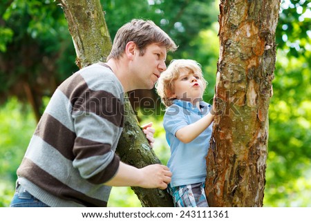 Father and his little cute son having fun in forest. Funny kid boy climbing on tree. Family leisure outdoors, on warm summer day.