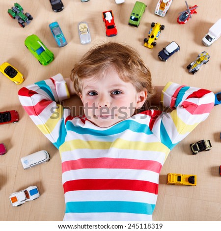 Little blond child playing with lots of toy cars indoor. Kid boy wearing colorful shirt and having fun.