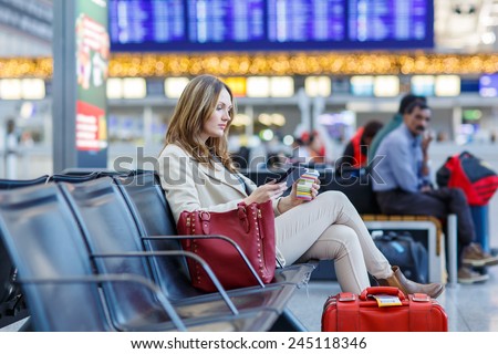 Young woman at international airport, reading her ebook computer and drinking coffee to go while waiting for her flight. Female passenger at terminal, indoors.