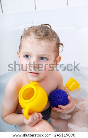 Adorable  blond toddler boy having fun with water by taking bath in bathtub at home. Funny kid splashing and playing with toys.