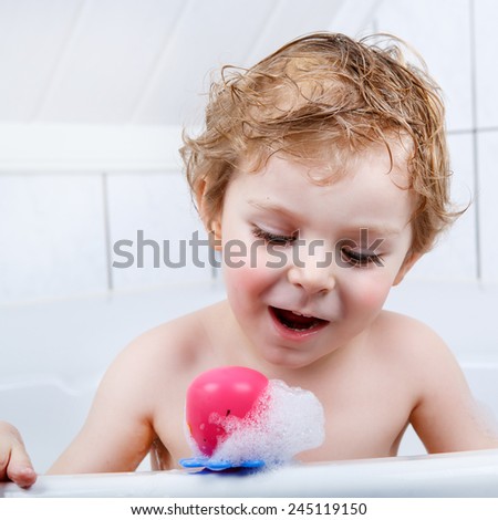 Adorable  blond toddler boy having fun with water by taking bath in bathtub at home. Funny kid splashing and playing with toys and foam.