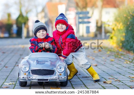 Two happy sibling boys in red jackets and rain boots playing with big old toy car, outdoors.  Kids leisure on cold day in winter, autumn or spring.