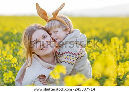 Little toddler child and his mother in Easter bunny ears having fun, celebrating traditional Easter holiday. In yellow rape field, outdoors, on warm spring day.