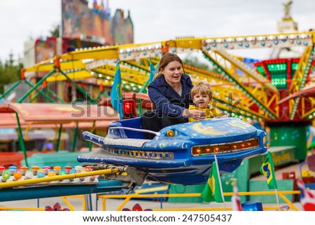 Happy mother and little son riding on a merry-go-round carousel together, smiling and having fun at a fair or amusement park. Active family leisure with kids.