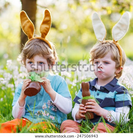 Family of two siblings: Little boys wearing Easter bunny ears at spring green grass and blooming apple garden, eating chocolate bunny and having fun outdoors.