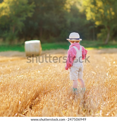 Funny little toddler boy in traditional German bavarian clothes, leather shorts and check shirt,  walking happily through wheat field near  hay stack bale. Active leisure with children on summer day.
