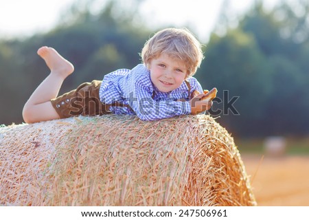Funny little kid boy in traditional German bavarian clothes, leather shorts and check shirt, lying on hay stack or bale and dreaming. Active outdoors leisure with children on warm summer day.