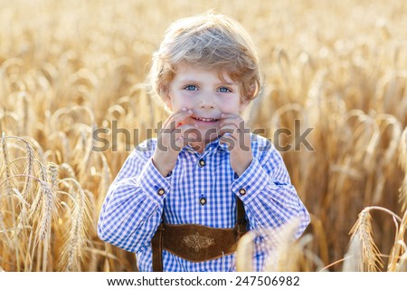 Funny little kid boy in traditional German bavarian clothes, leather shorts and check shirt, walking happily through wheat field near  hay stack or bale. Active leisure with children on summer day.