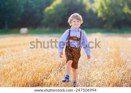 Funny little kid boy in traditional German bavarian clothes, leather shorts and check shirt, walking happily through wheat field near  hay stack  bale. Active leisure with children on warm summer day