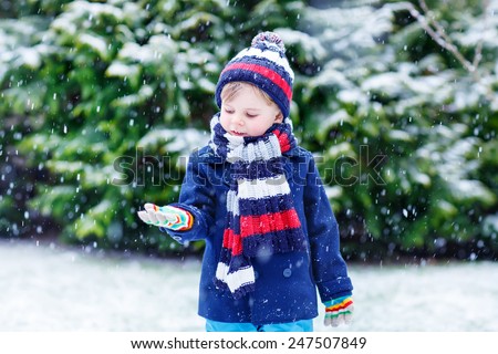 Cute little funny boy in colorful winter clothes catching snow and snowflakes, outdoors during snowfall. Active outdoors leisure with children in winter. Kid with warm hat, hand gloves and scarf