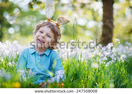Cute happy little boy wearing Easter bunny ears at spring green grass and blooming apple garden, lauging and having fun outdoors.