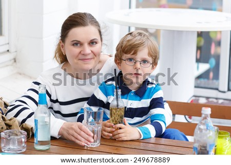 Little cute boy with glasses and his mother enjoying drinks in outside cafe in summer. Happy family having fun together.