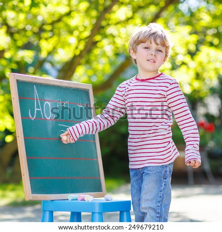 Little blond child at blackboard practicing writing letters and mathematics, outdoor school or nursery. Kid learning and schoolboy concept. On summer sunny day. Back to school.