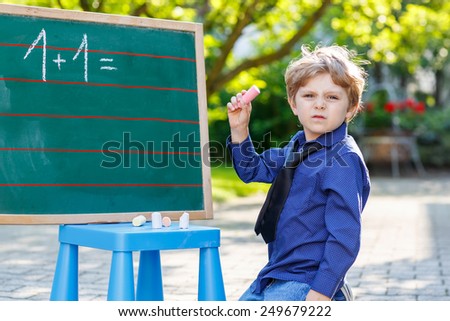Little confused boy at blackboard practicing writing letters and mathematics, outdoor school or nursery. Kid learning and schoolboy concept. On summer sunny day.