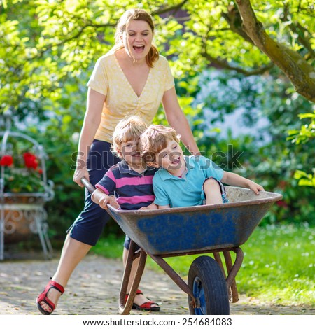 Two little twins having fun in a wheelbarrow pushing by mum  in domestic garden, on warm sunny day. Active outdoors games for kids in summer.