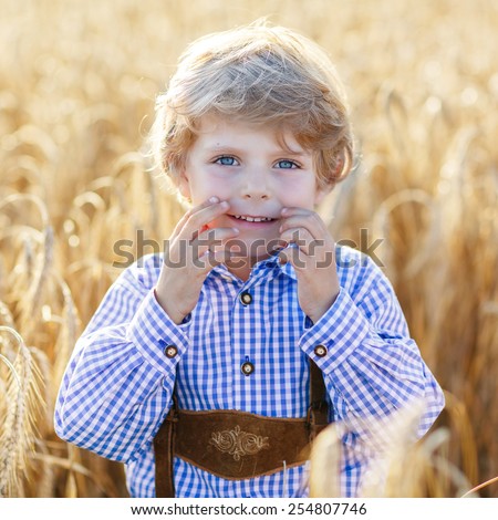 Funny little kid boy in traditional German bavarian clothes, leather shorts and check shirt, walking happily through wheat field near  hay stack or bale.