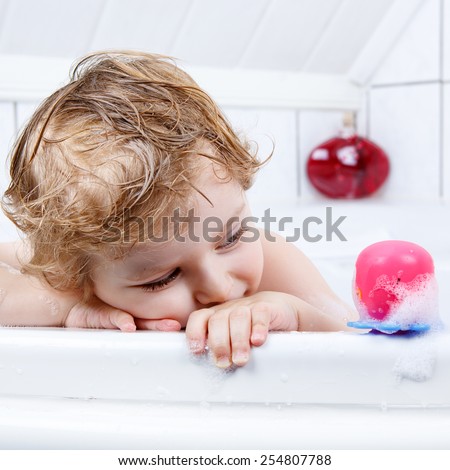 Happy cute little  blond toddler boy having fun with water by taking bath in bathtub at home. Funny kid splashing and playing with toys.