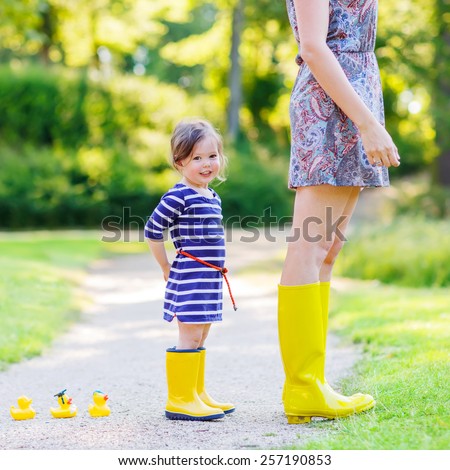 Woman and her little cute kid daughter in yellow rubber boots, family look, playing with duck toys in summer park. Active leisure with kids.