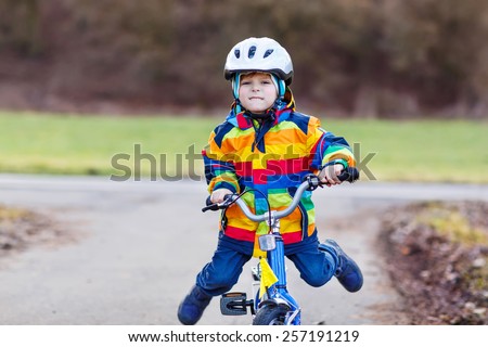 Funny cute  preschool kid boy in safety helmet and colorful raincoat riding his first bike and having fun on cold  day, outdoors. Active leisure with children in winter, spring or autumn.