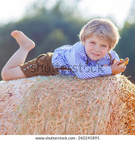 Funny little kid boy in traditional German bavarian clothes, leather shorts and check shirt, lying on hay stack or bale and dreaming. Active outdoors leisure with children on warm summer day.