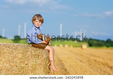 Funny little kid boy in traditional German bavarian clothes, leather shorts and check shirt, sitting on hay stack or bale and eating pretzel. Active outdoors leisure with children on warm summer day.