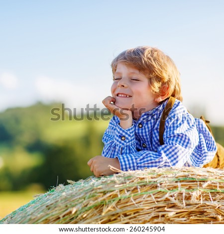 Happy little kid boy in traditional German bavarian clothes, leather shorts and check shirt,  lying on hay stack or bale and dreaming. Active outdoors leisure with children on warm summer day.